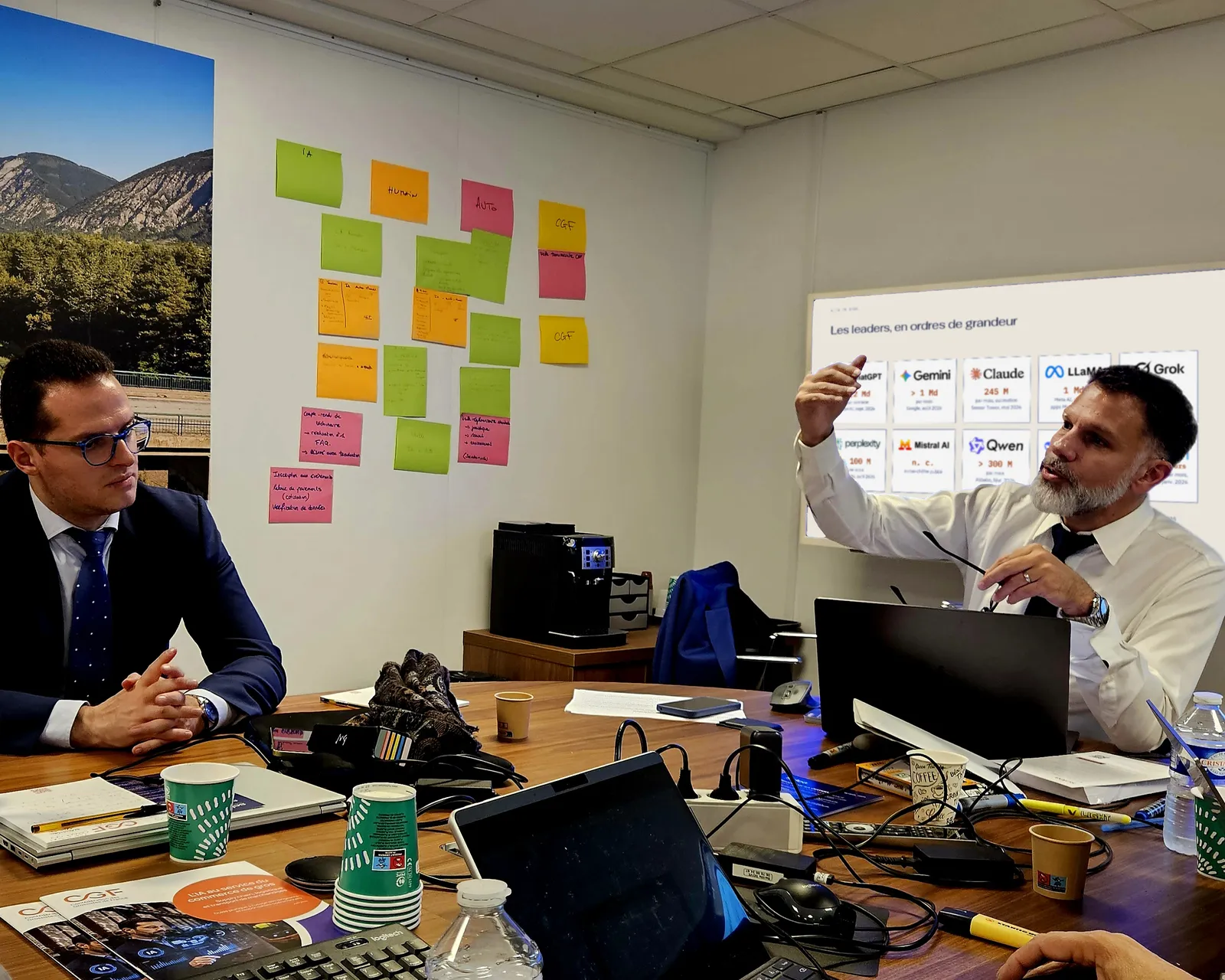 Séance de travail autour de la table de réunion de la CGF : à droite, le formateur explique en désignant l’écran, où s’affiche une diapositive sur les principaux assistants d’IA ; à gauche, un participant l’écoute.