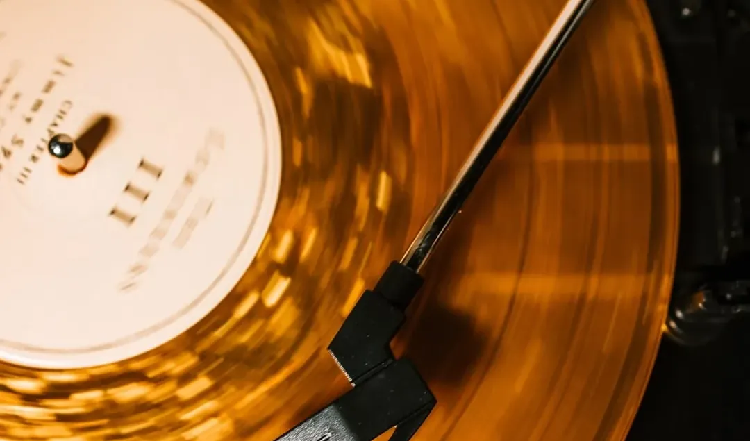 Vue de dessus d’un disque vinyle ambré en rotation sur une platine, le mouvement rendant les sillons flous. Le bras de lecture traverse l’image en diagonale, sa cellule posée dans le sillon.