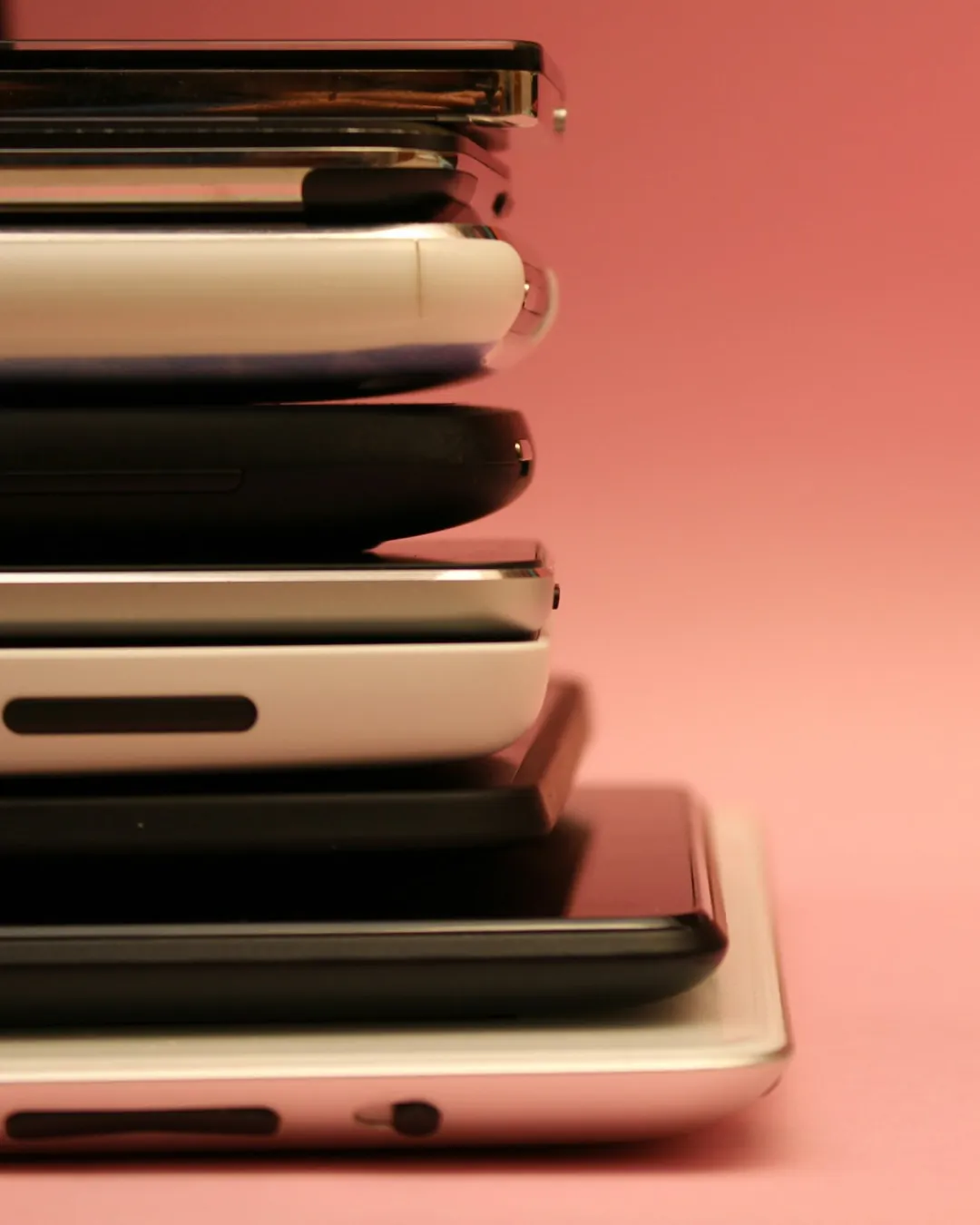 Pile de smartphones et de tablettes posés à plat les uns sur les autres et vus de profil, du plus petit en haut au plus grand en bas, boîtiers blancs et noirs alternés, sur un fond uni rose.