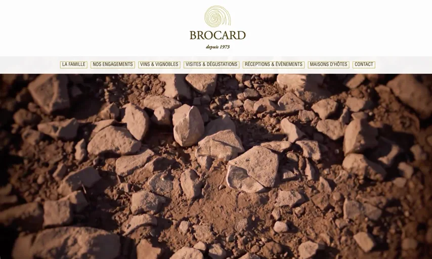 Page d'accueil du site du Domaine Jean-Marc Brocard : le logo Brocard « depuis 1973 » au-dessus d'un menu portant La famille, Nos engagements, Vins & vignobles, Visites & dégustations, Réceptions & évènements, Maisons d'hôtes et Contact, puis une photographie plein cadre d'un sol de vigne caillouteux.
