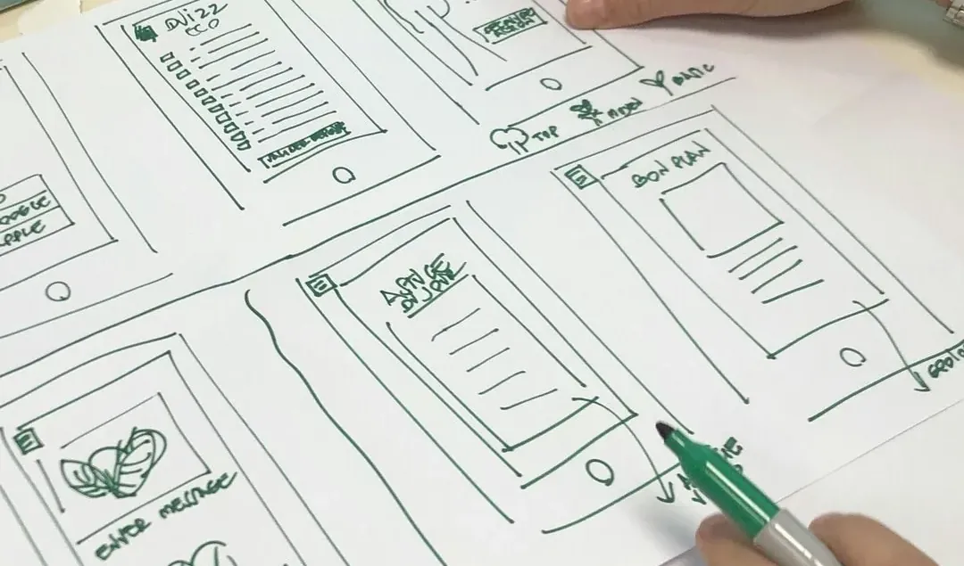 Planche de croquis d’écrans mobiles sur une table d’atelier, deux mains au travail, l’une pointant un écran, l’autre tenant un feutre, post-its en arrière-plan.