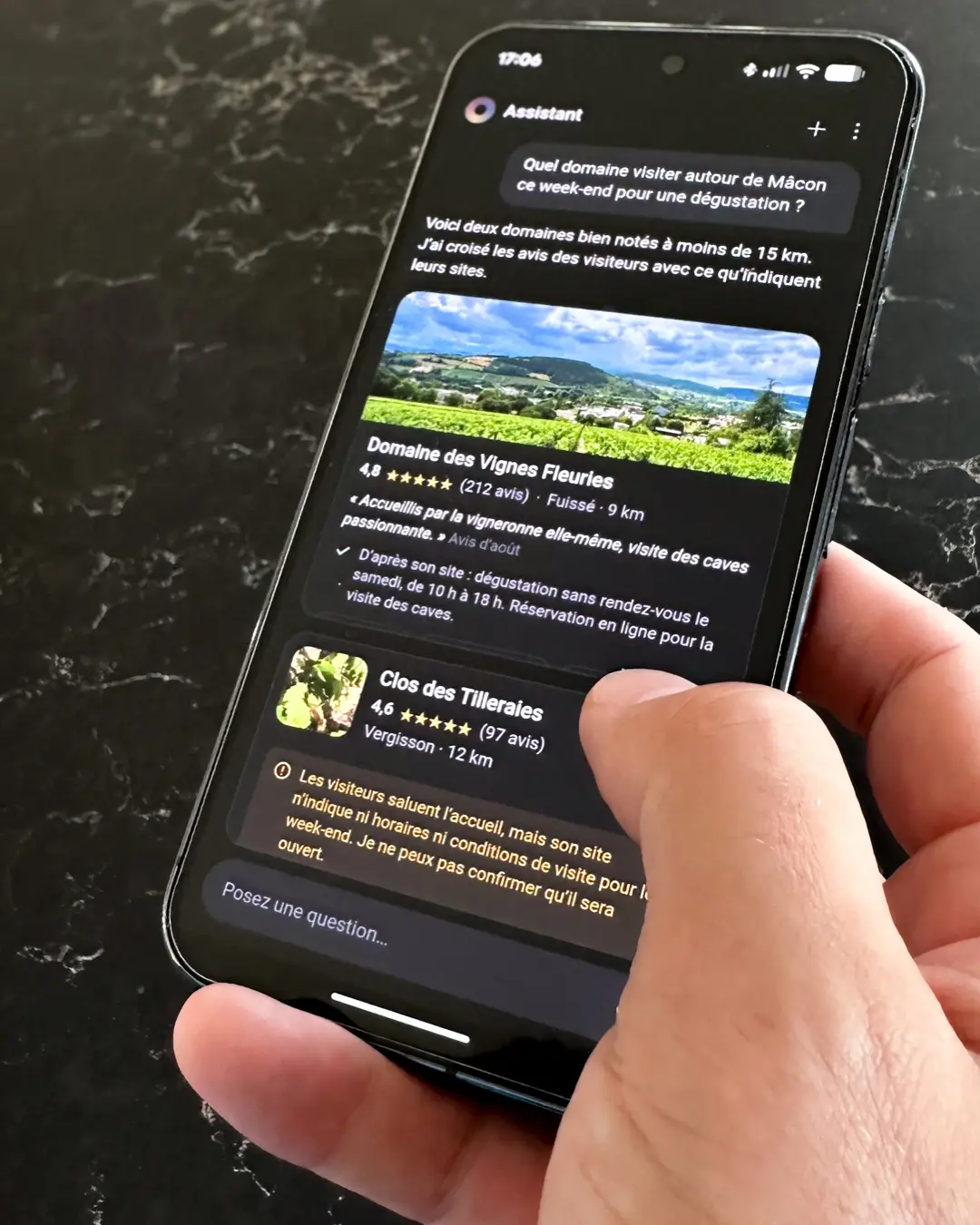 Main tenant un téléphone qui affiche un assistant conversationnel. À la question « Quel domaine visiter autour de Mâcon ce week-end pour une dégustation ? », l’assistant propose deux domaines bien notés à moins de 15 km. Pour le premier, il confirme d’après son site la dégustation sans rendez-vous le samedi. Pour le second, il signale que le site n’indique ni horaires ni conditions de visite, et qu’il ne peut donc pas confirmer qu’il sera ouvert. Écran fictif réalisé pour l’illustration, domaines imaginaires.